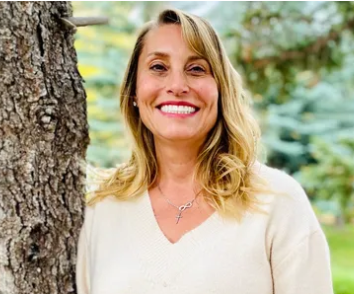 A smiling person with blonde hair wearing a light sweater and an infinity necklace, standing outdoors by a tree.