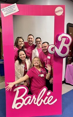 A group of six people posing behind a Barbie-themed cardboard photo frame, smiling and wearing shades of pink.
