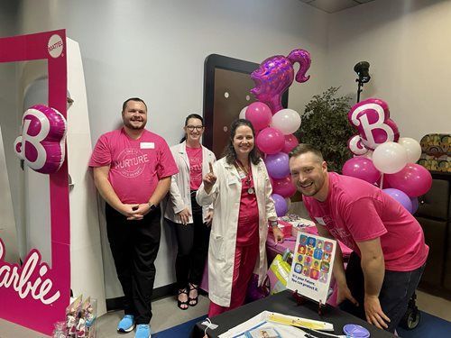 Four people in pink and white attire pose by a Barbie-themed display with pink balloons and decorations.
