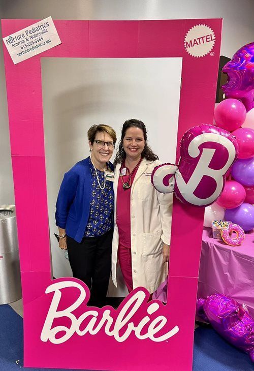 Two smiling people stand behind a pink Barbie-themed photo frame surrounded by pink balloons at a pediatric office event.