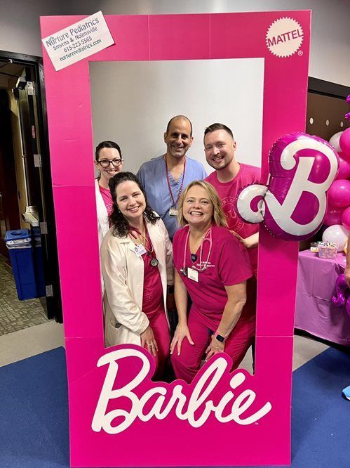 Five healthcare workers in scrubs and coats pose behind a large, pink cardboard Barbie box frame at an event.