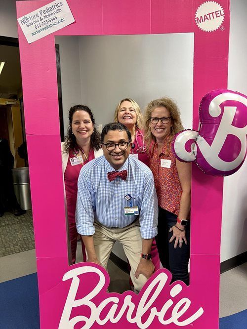 Four smiling people pose inside a large pink Barbie-themed photo frame in an office setting.