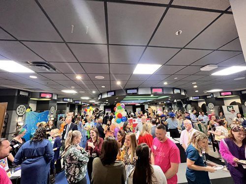 People socialize at a crowded networking event in a modern room with a dark ceiling and rows of television screens.
