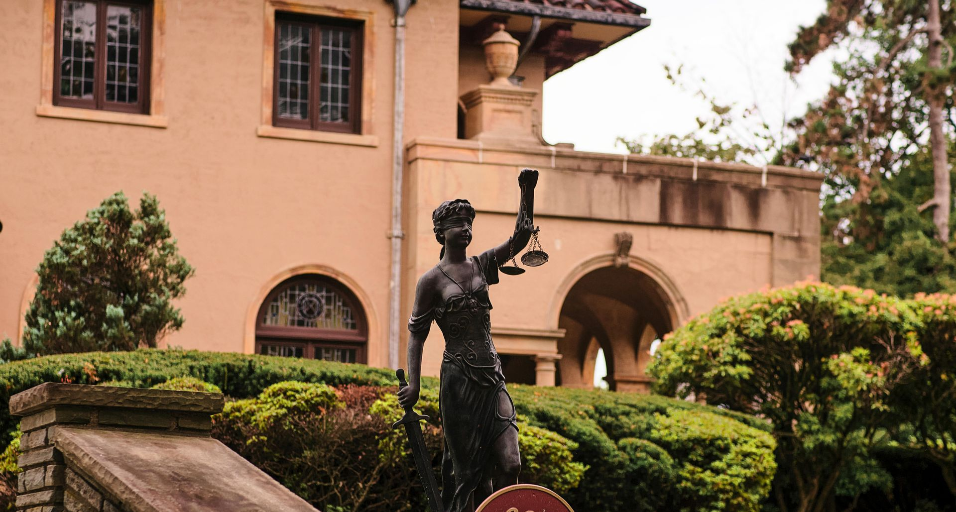 Estátua de bronze em um pátio ajardinado em frente a um edifício cor de pêssego com janelas em arco e sebes.