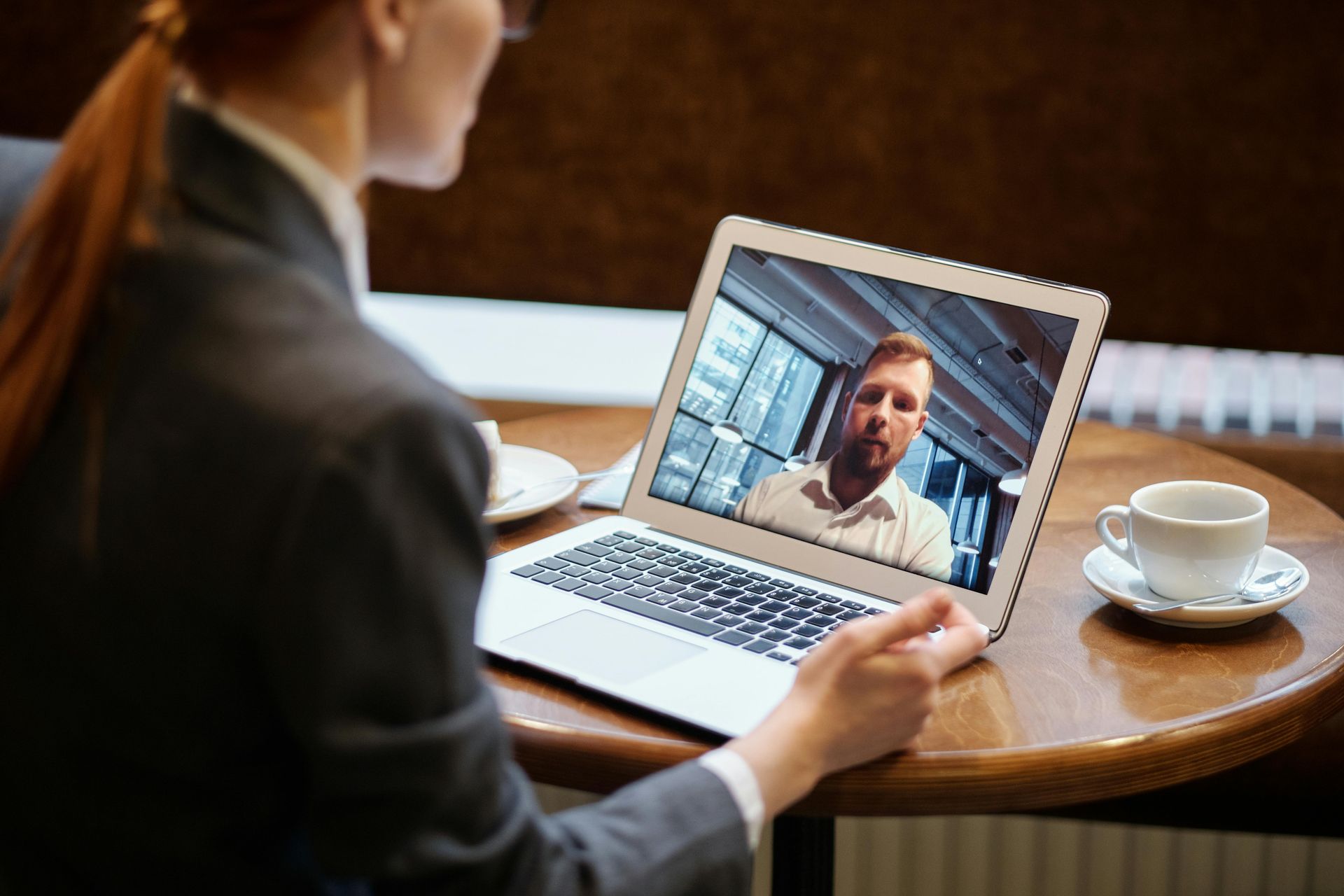 Pessoa em chamada de vídeo com um homem usando um laptop em uma mesa de café, ao lado de uma xícara de café.