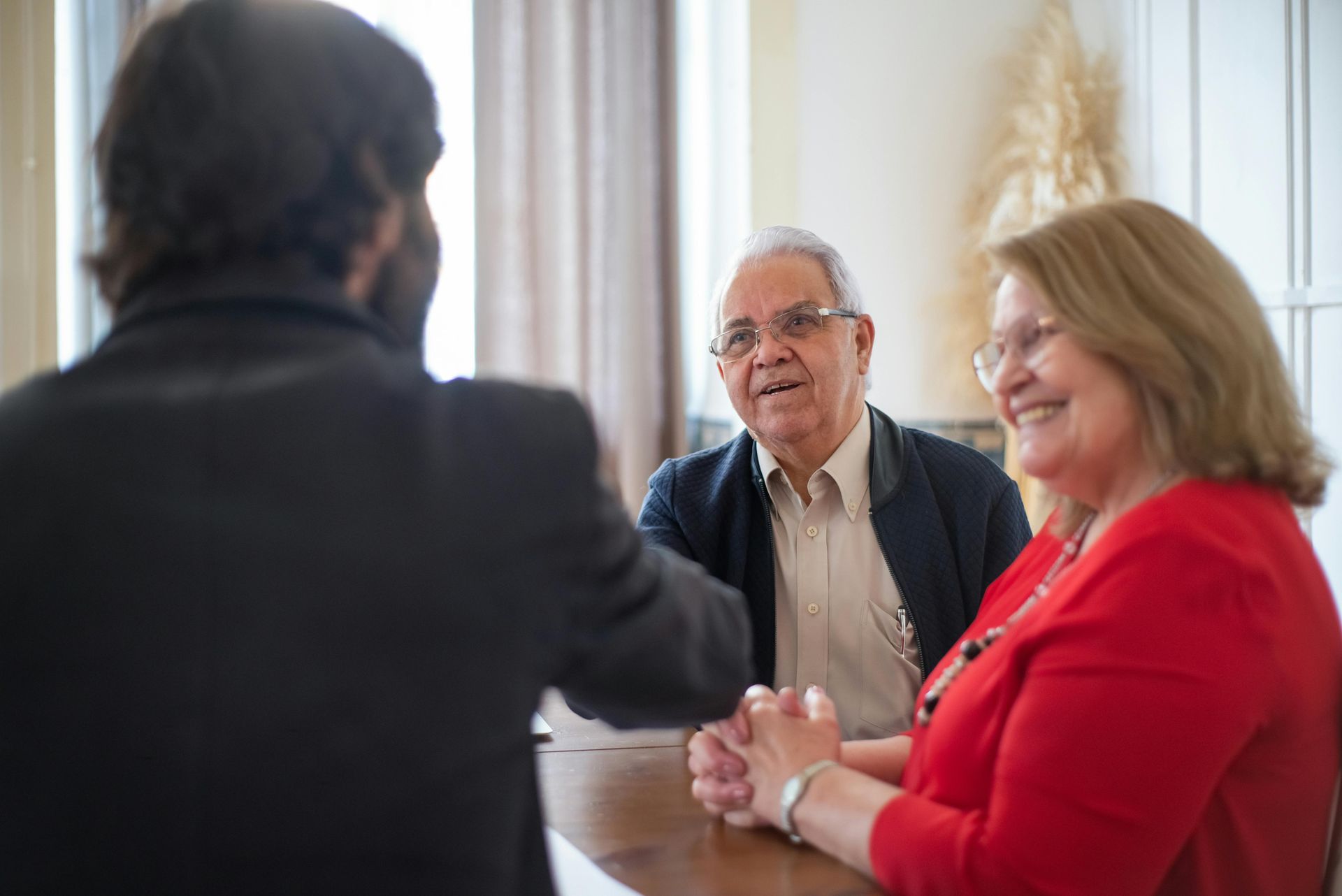 Três pessoas conversando à mesa, com um homem sorrindo ao centro e uma mulher de vermelho à direita.