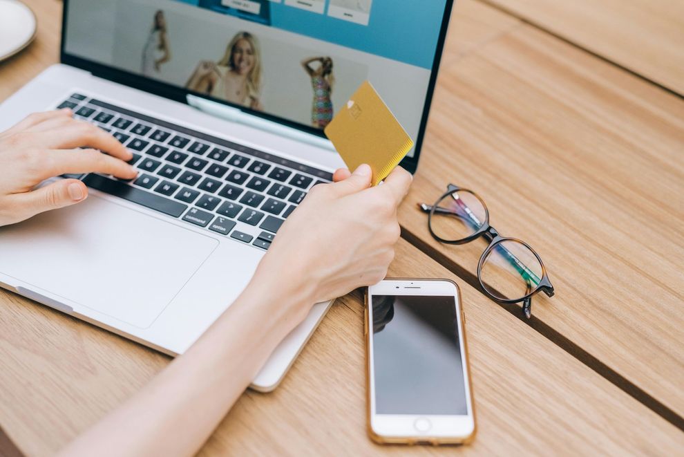 Mãos usando laptop e cartão em uma mesa com telefone e óculos por perto