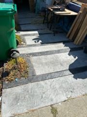 Steps leading to a doorway, damaged concrete with black trim. A green trash can and a plant are on the left.
