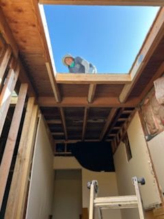 Person leans out of a skylight during construction. Framing and ladder visible. Bright blue sky.