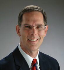 A woman in a white shirt with flowers on it is smiling in front of a blue background
