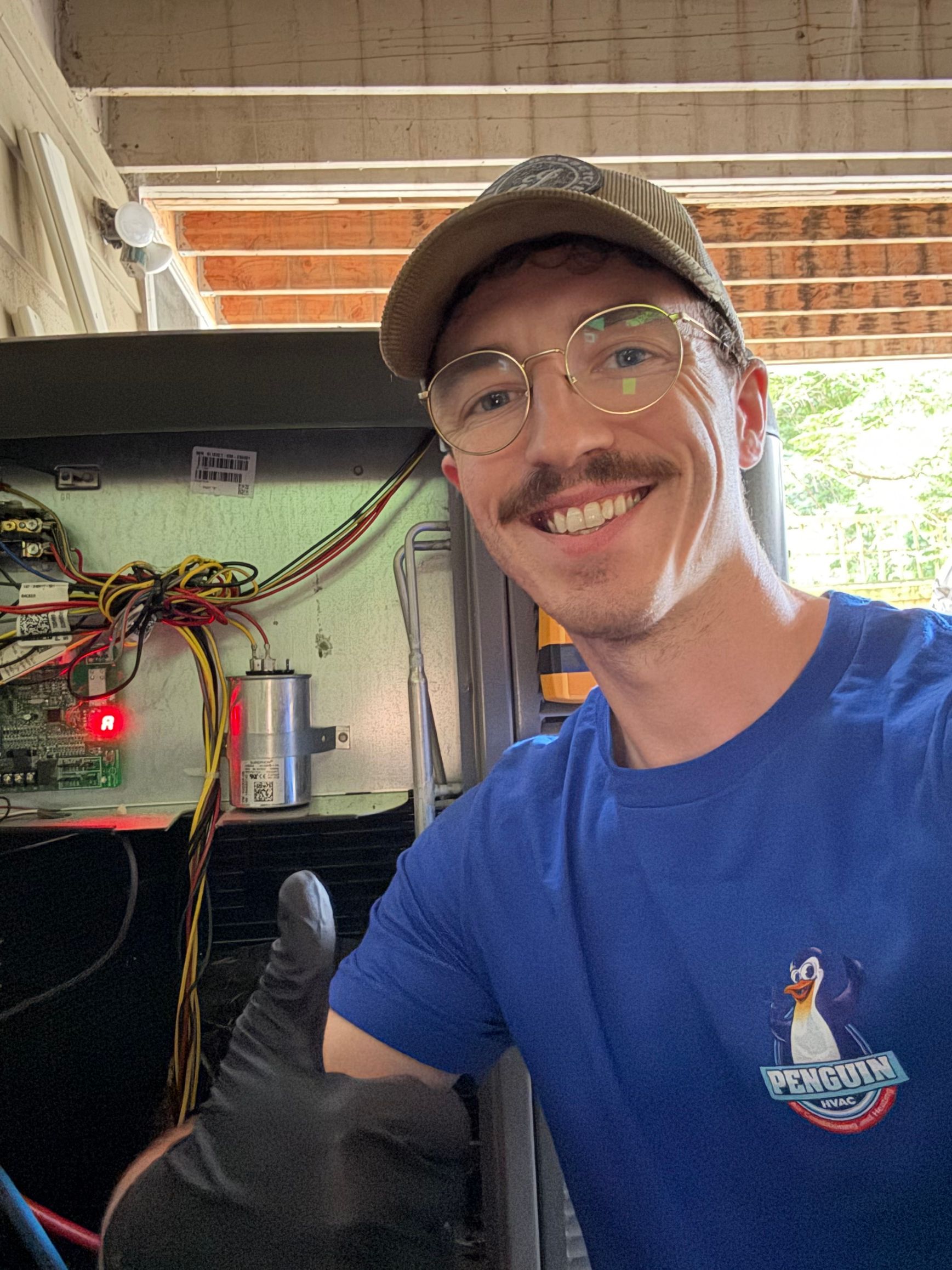 Man in blue shirt and baseball cap with thumbs up, working on electrical panel, smiling.