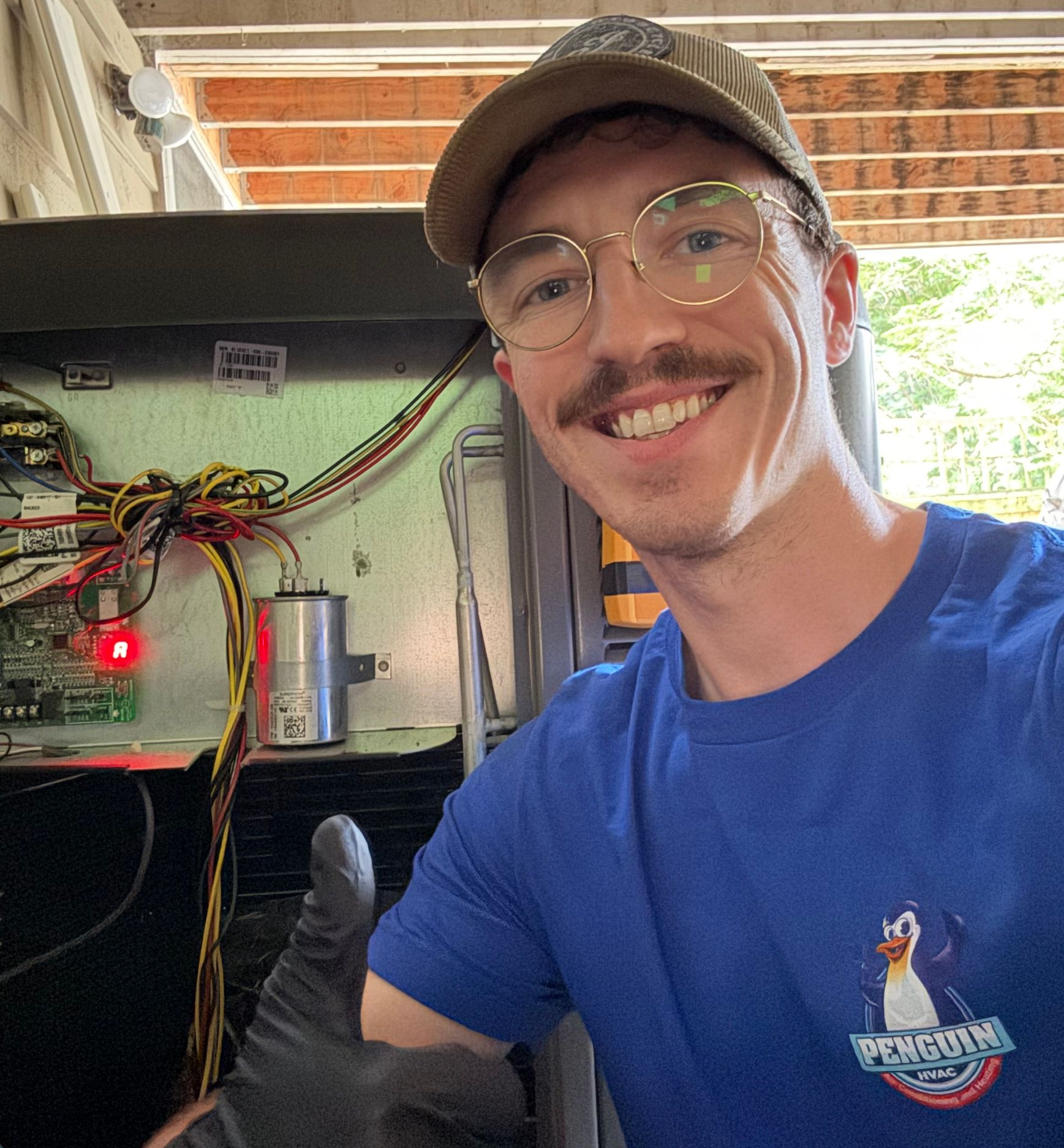 Man in cap and glasses gives thumbs up next to machinery. Wears blue shirt with penguin logo.