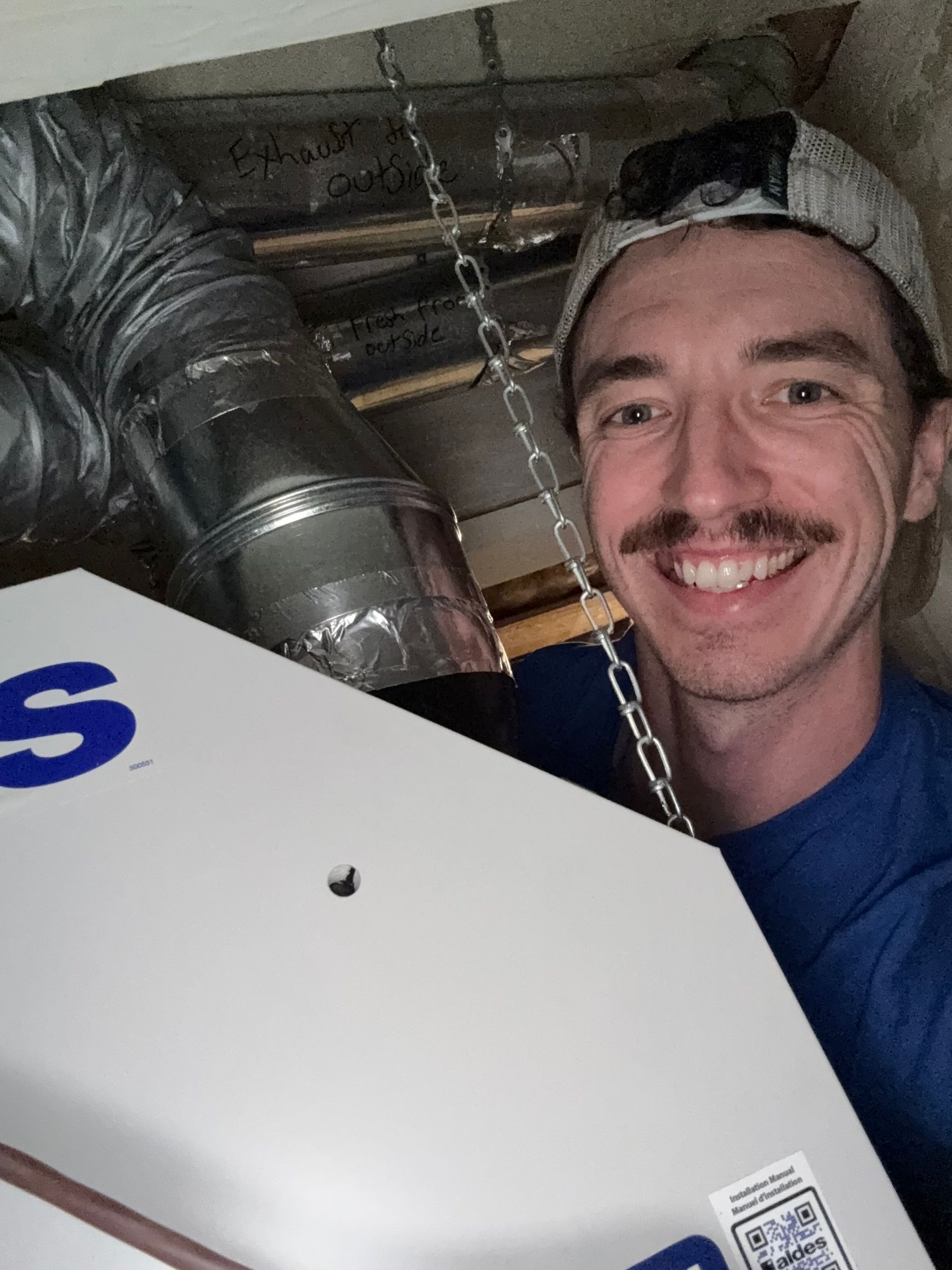 Man smiles, holding HVAC unit. Silver ductwork and ceiling visible.