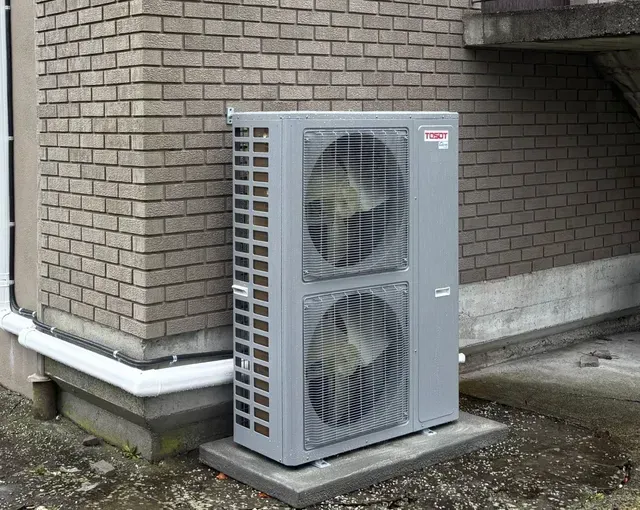 A grey, dual-fan outdoor heat pump unit mounted on a concrete pad against a brick exterior wall.