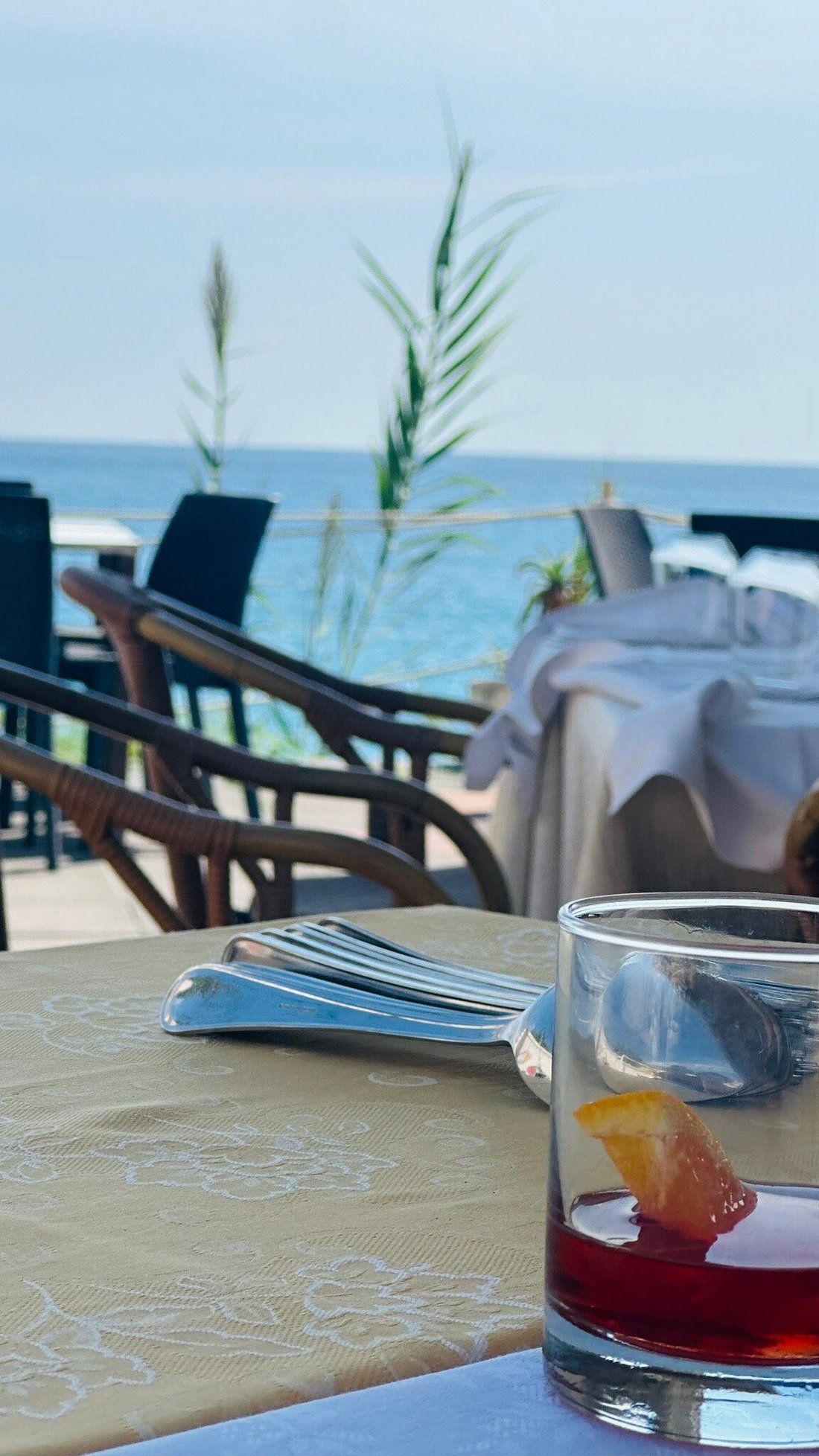 Tavolino sulla spiaggia con un bicchiere di bevanda rossa, tovaglioli piegati e sedie con vista sul mare.