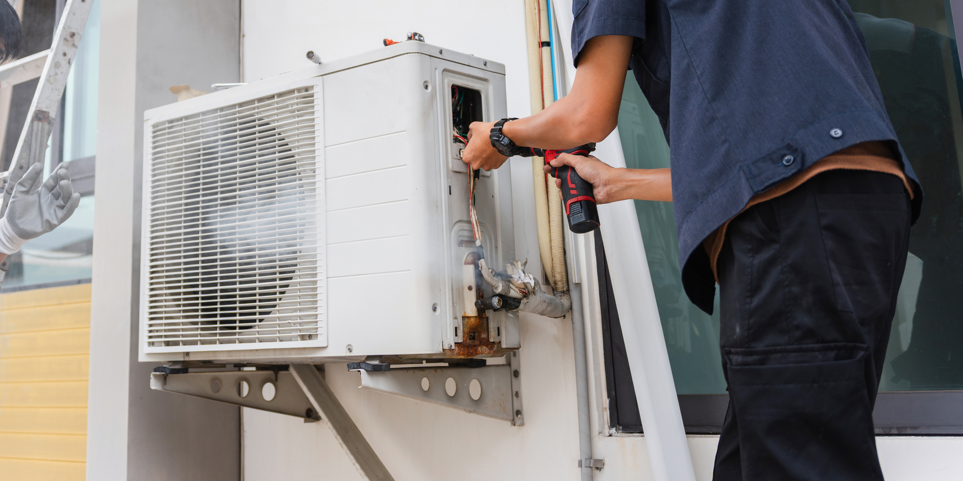 Technician in a blue uniform using a drill to repair an outdoor air conditioning unit mounted on a wall.