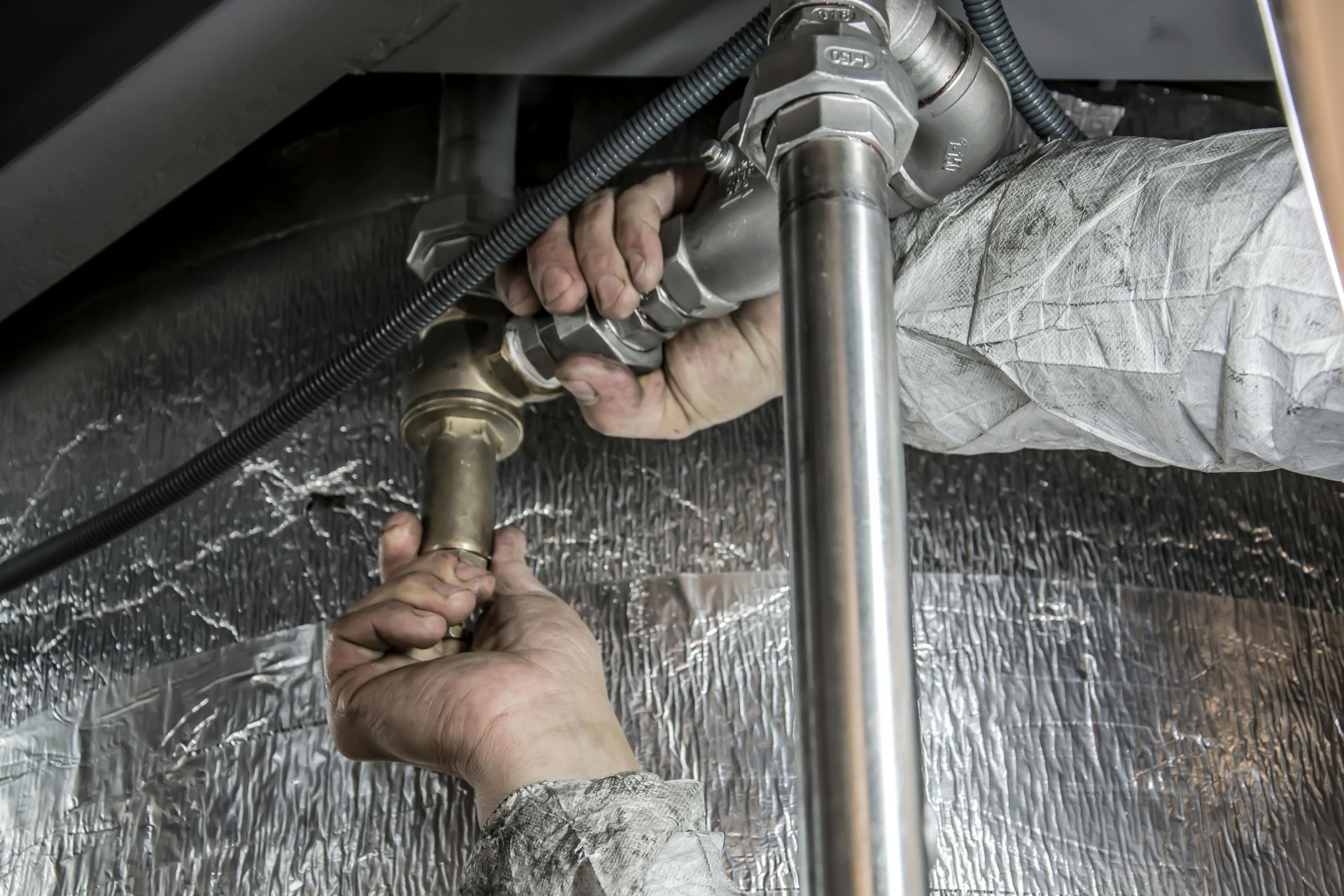 A person in work coveralls uses both hands to tighten a metallic pipe fitting against a reflective insulation background.