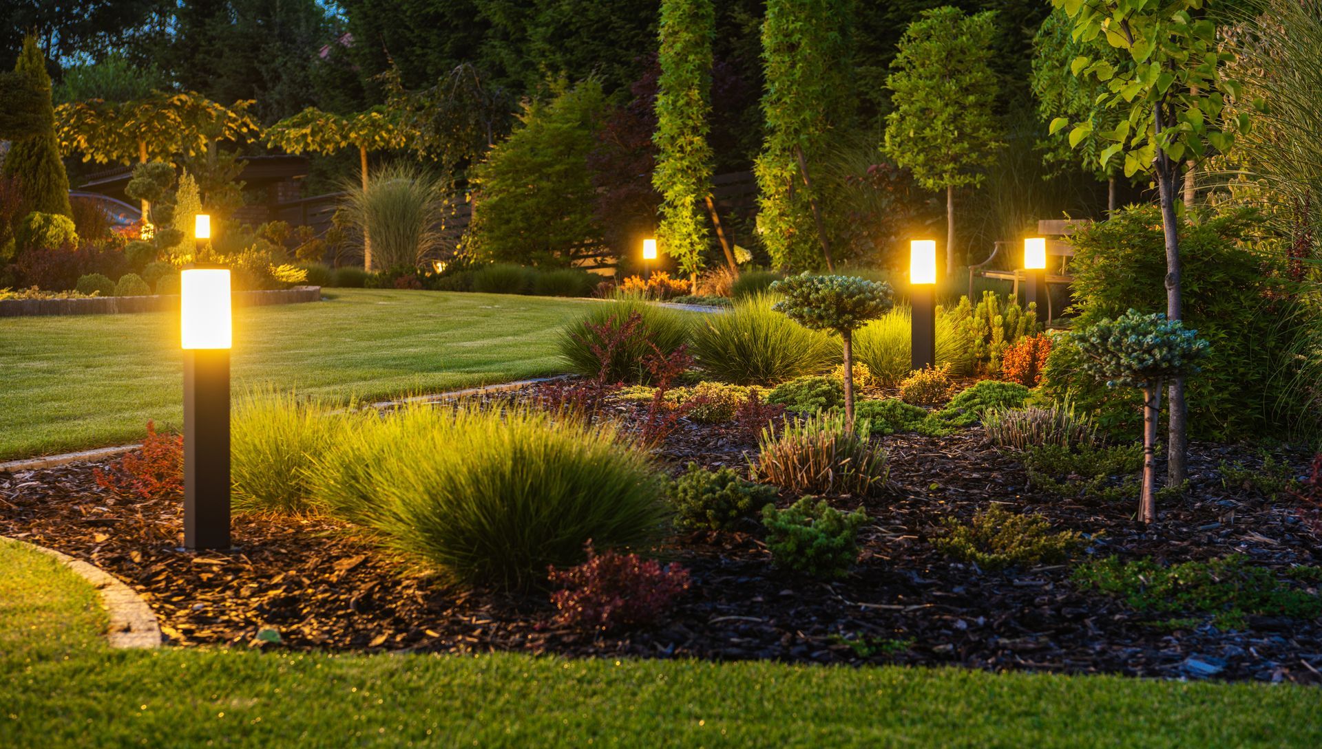 Illuminated garden path with glowing landscape lights, shrubs, flowers, and a manicured lawn at dusk