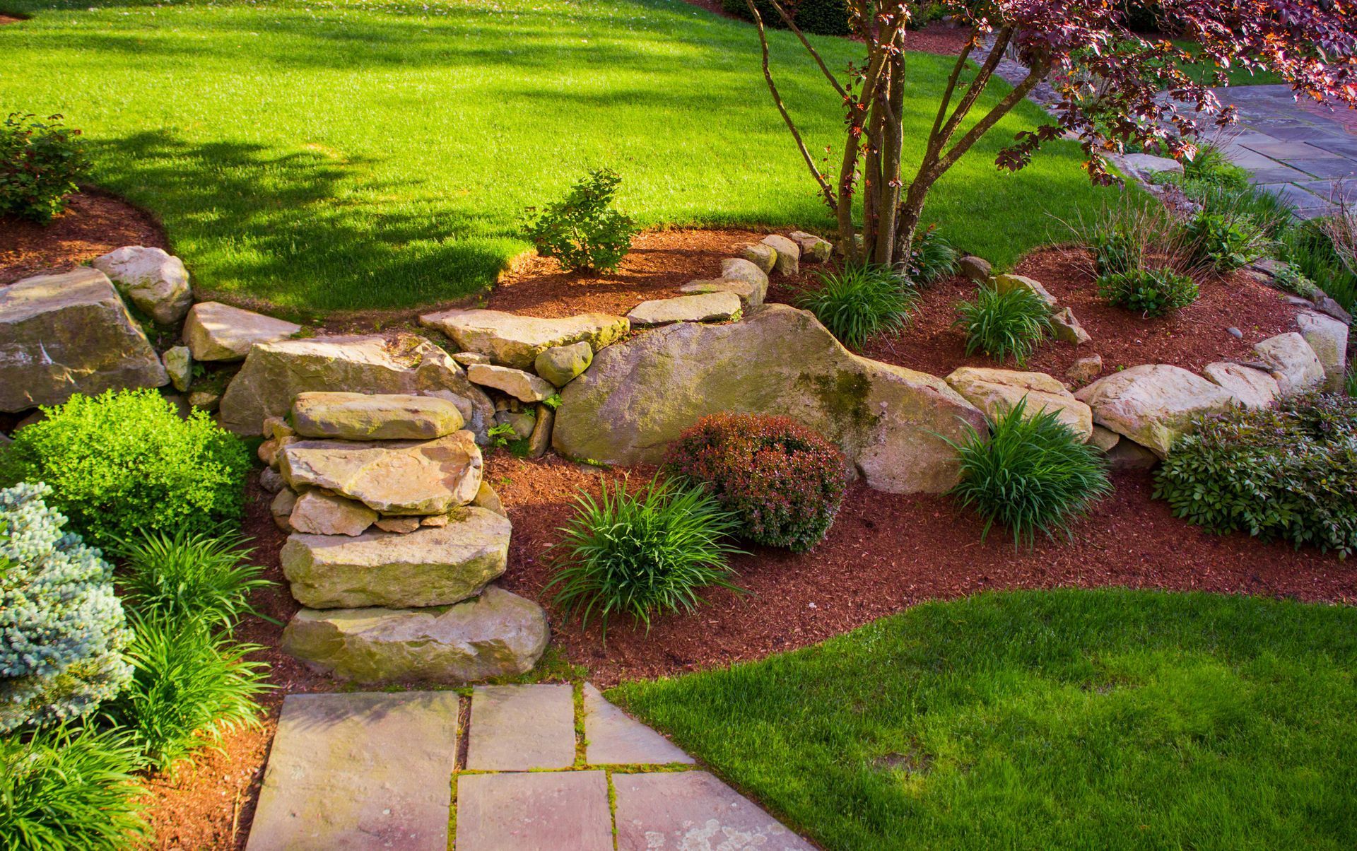 Stone garden border with stacked rocks, green shrubs, and a paved path beside a lawn.