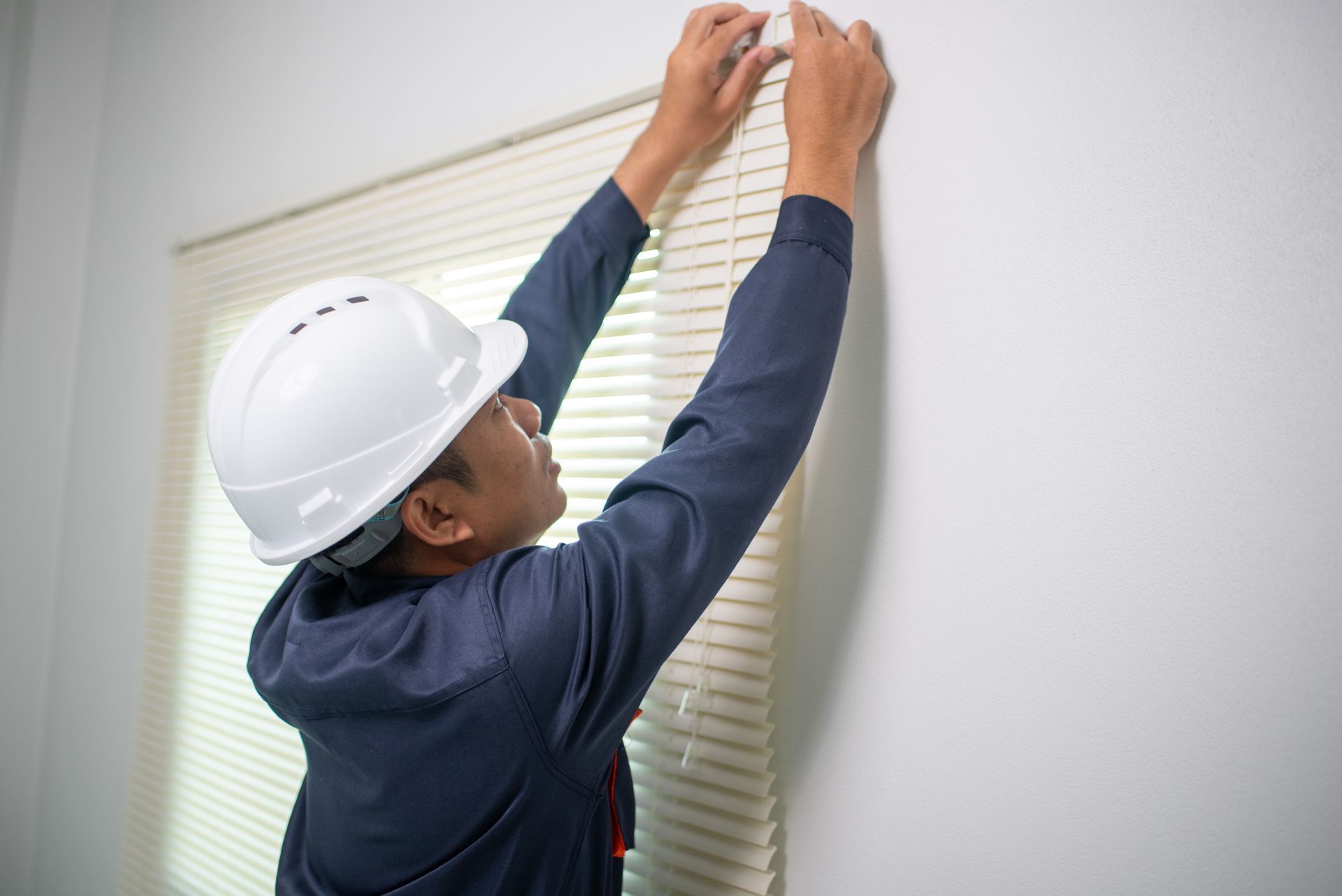 A man wearing a hard hat is hanging blinds on a wall.