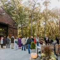 Guests gathered outside a rustic barn venue under autumn trees, mingling near tables and potted plants.