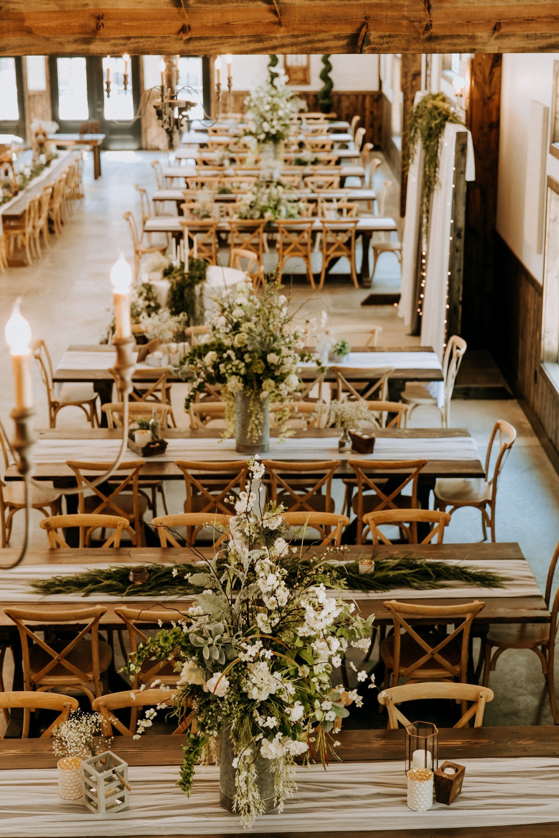 Rustic barn wedding reception setup with long wooden tables, cross-back chairs, and floral centerpieces