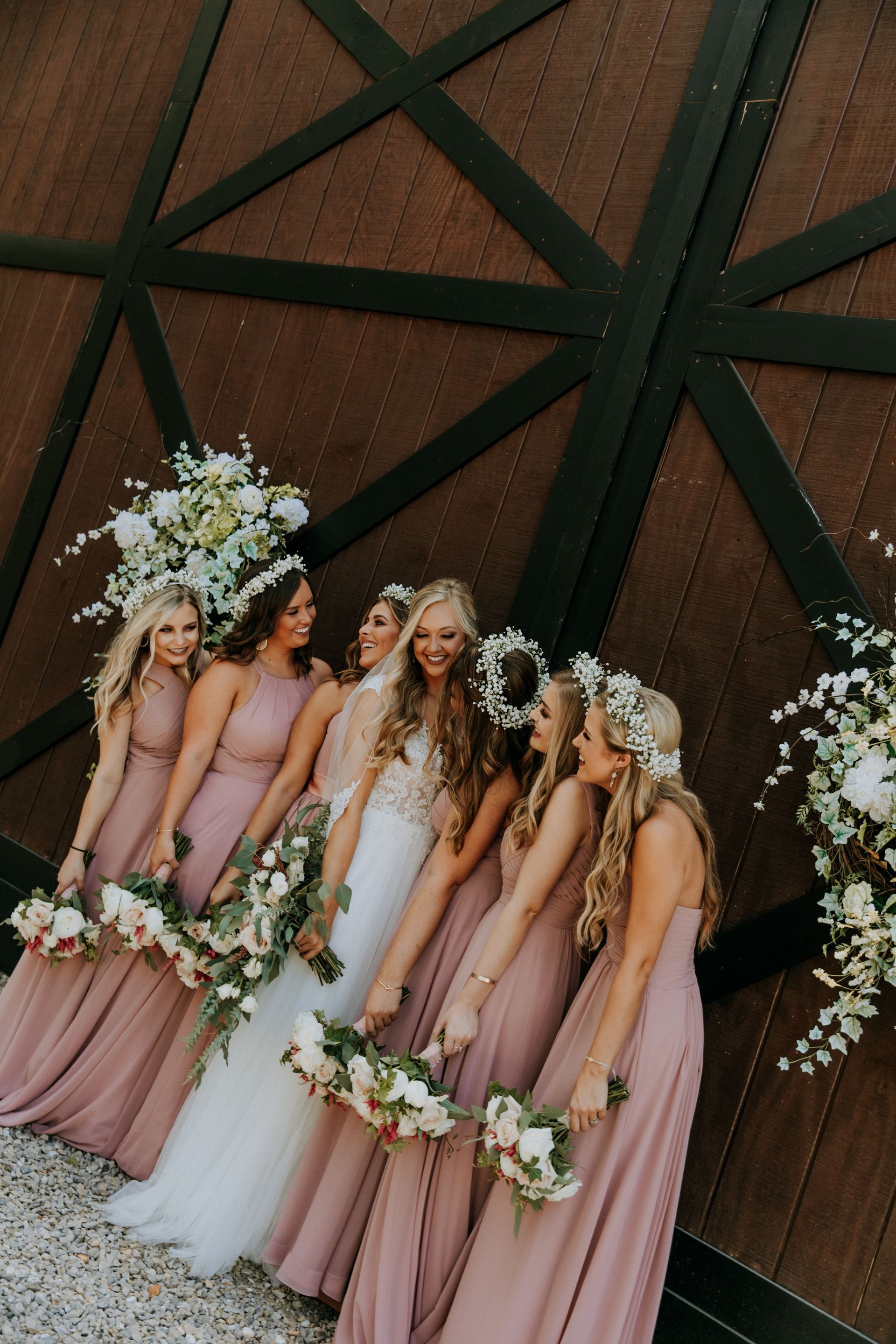 Five women in blush dresses and flower crowns holding white floral hoops in a rustic barn setting