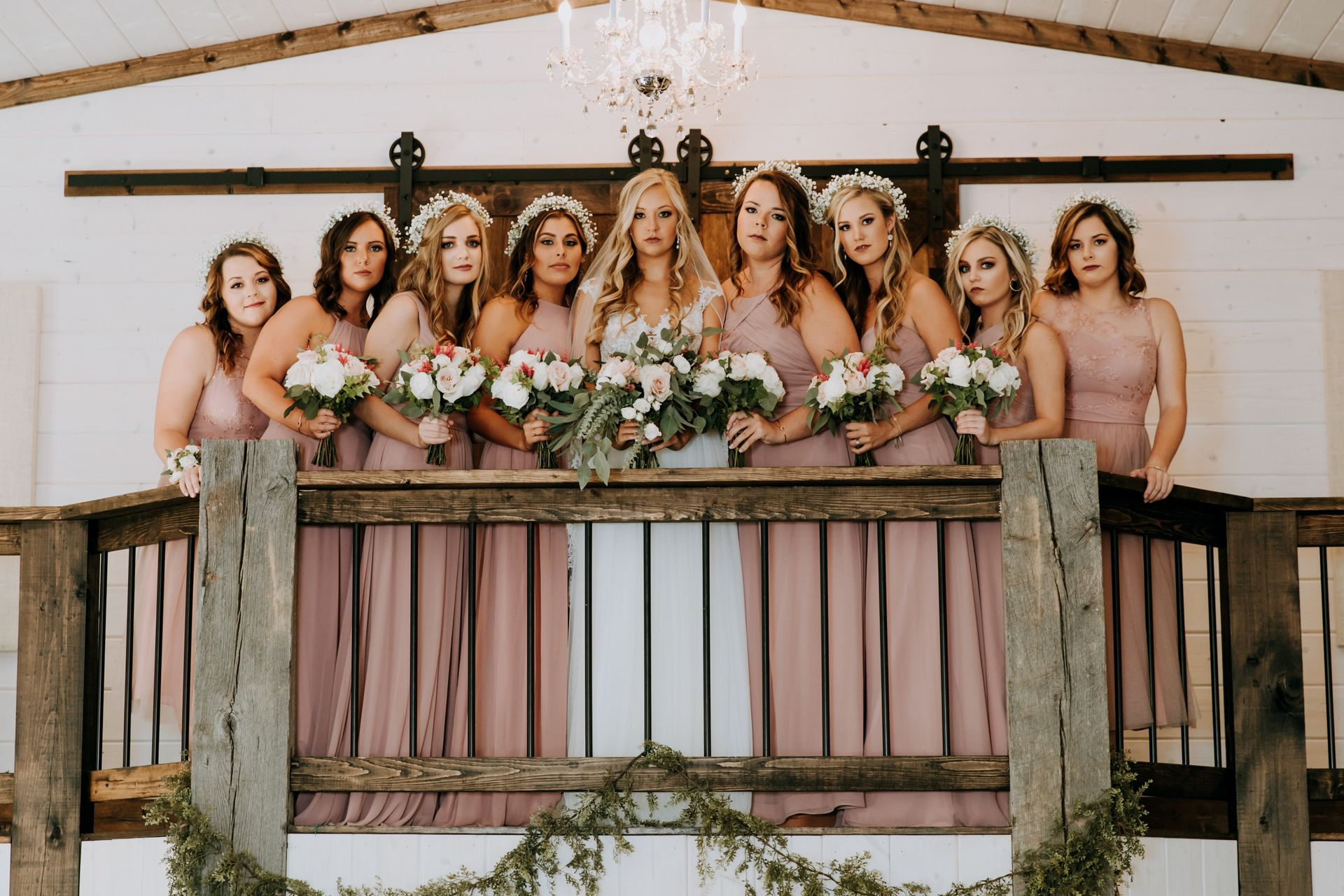 Group of bridesmaids in blush dresses holding bouquets on a decorated balcony indoors