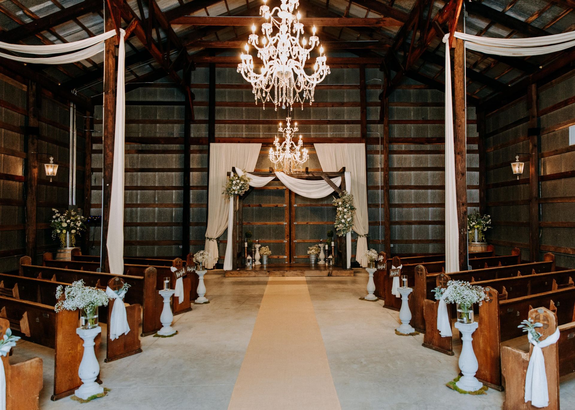 Rustic wedding ceremony setup in a barn with decorated aisle, chandeliers, and draped altar