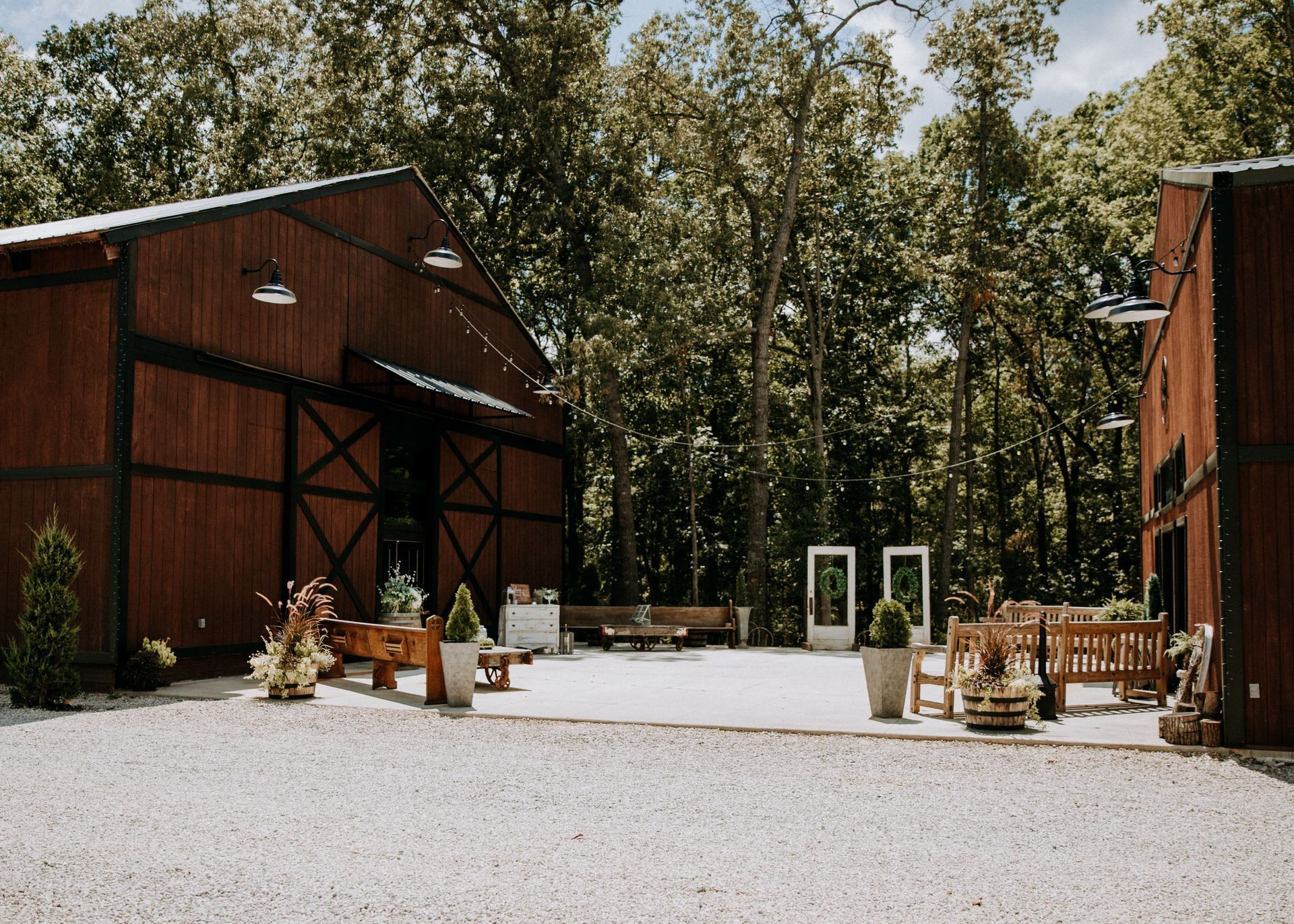 Rustic barn courtyard with red wooden buildings, gravel ground, and outdoor seating among trees
