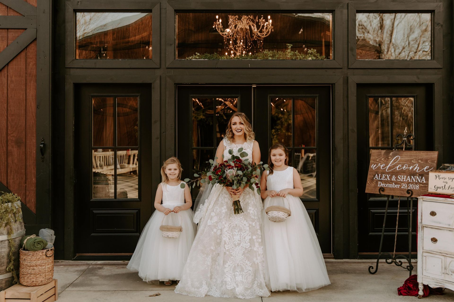 Bride in a white gown stands with two flower girls in white dresses outside a venue entrance