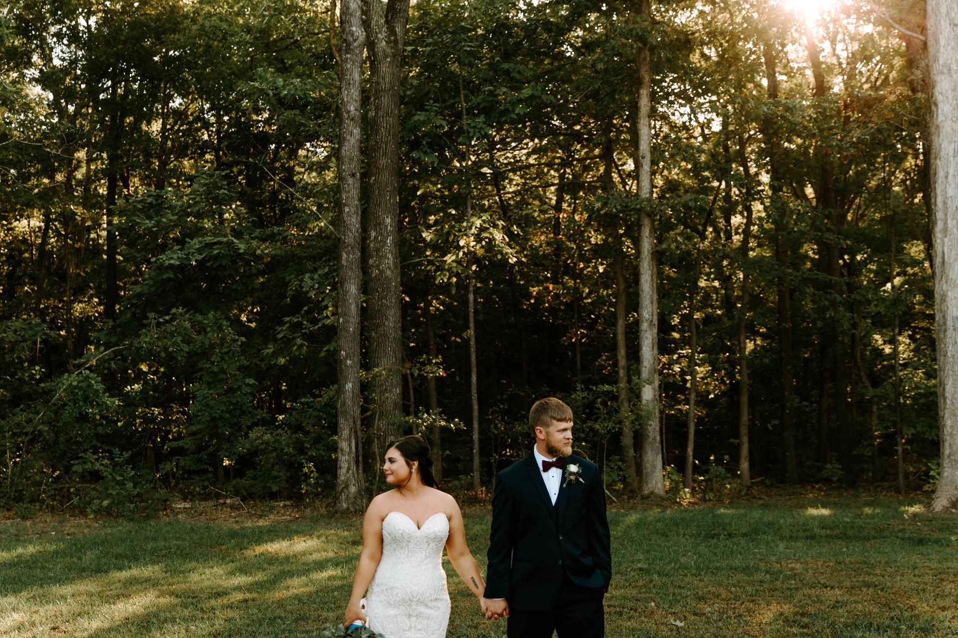 Bride and groom in wedding attire standing in a wooded clearing at sunset