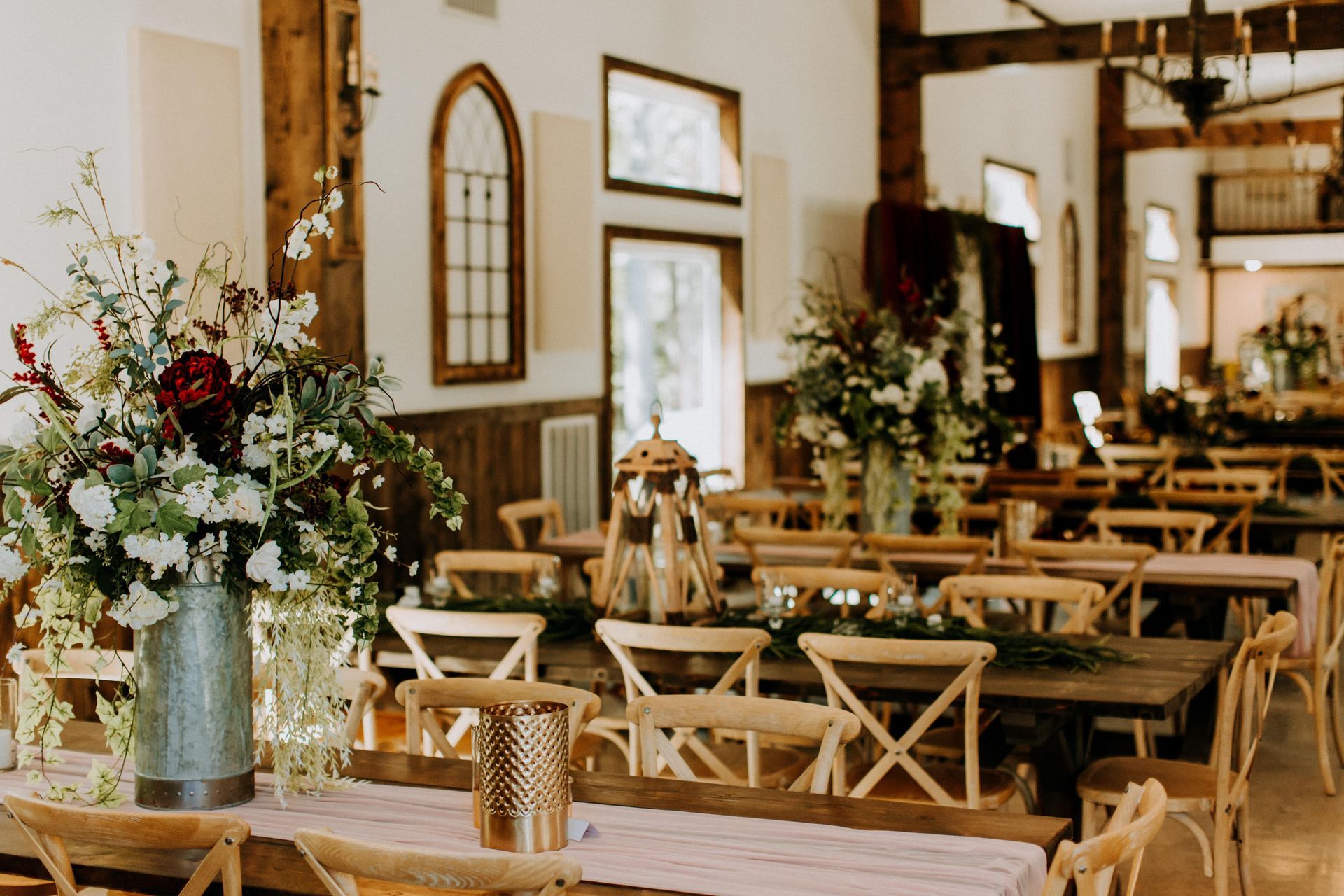 Rustic wedding reception hall with wooden tables, cross-back chairs, and floral centerpieces