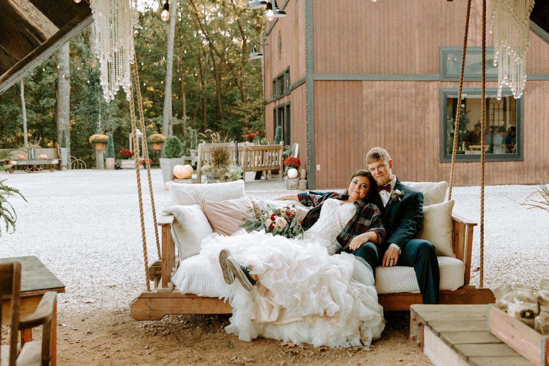 Two people lounging on a wooden outdoor daybed with white blankets in a rustic patio setting