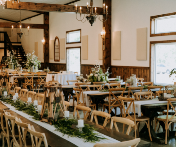 Rustic wedding reception hall with long banquet tables, greenery, candles, and wooden chairs