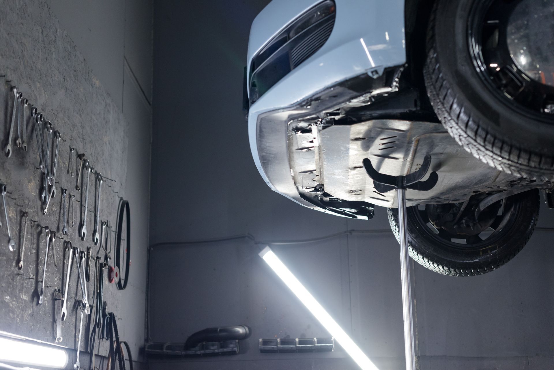 Car on a lift in a garage. Tools hang on the wall. A bright light shines beneath the vehicle.