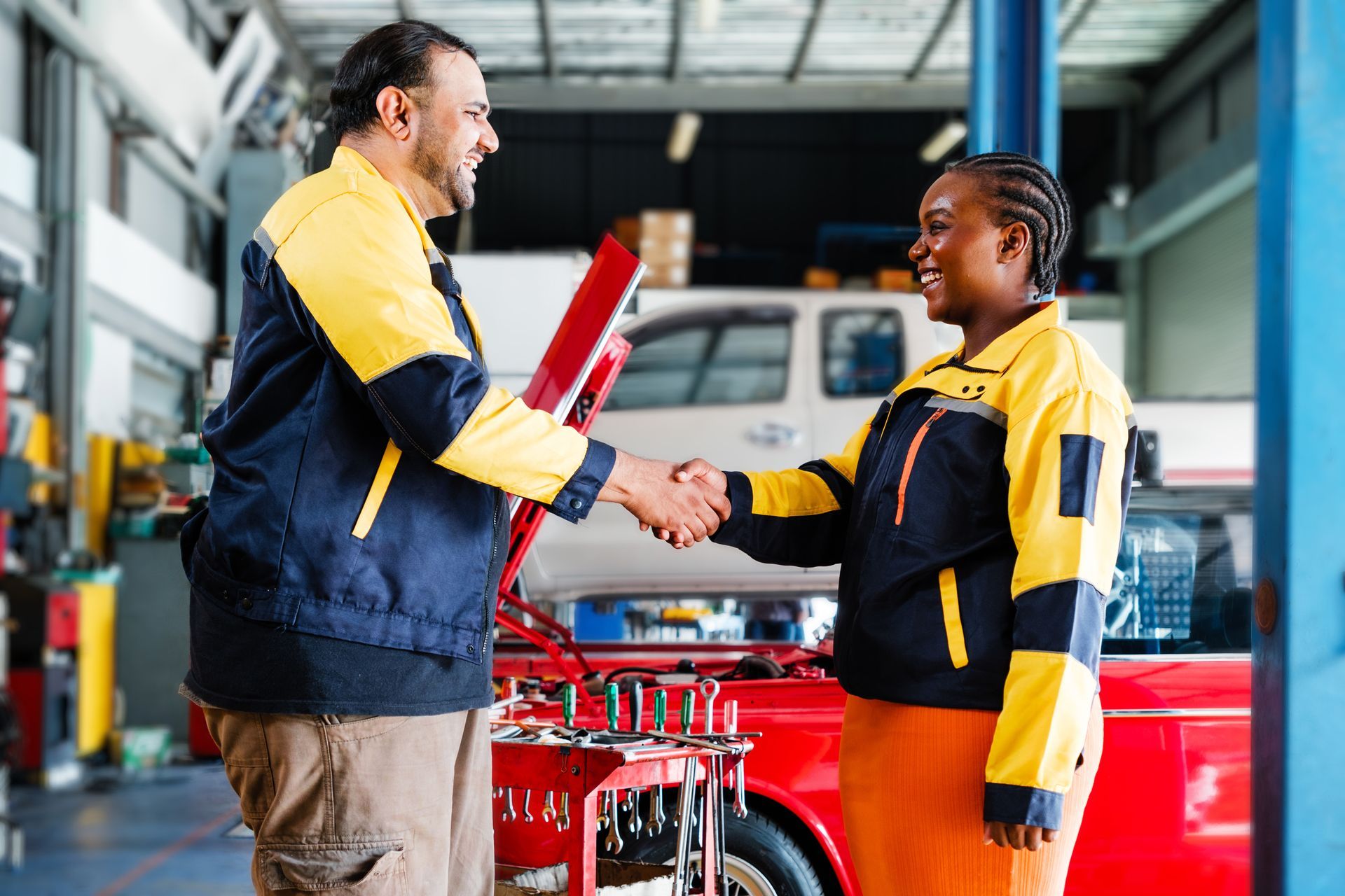 Two mechanics shaking hands in a car repair shop. Both wear navy and yellow uniforms.