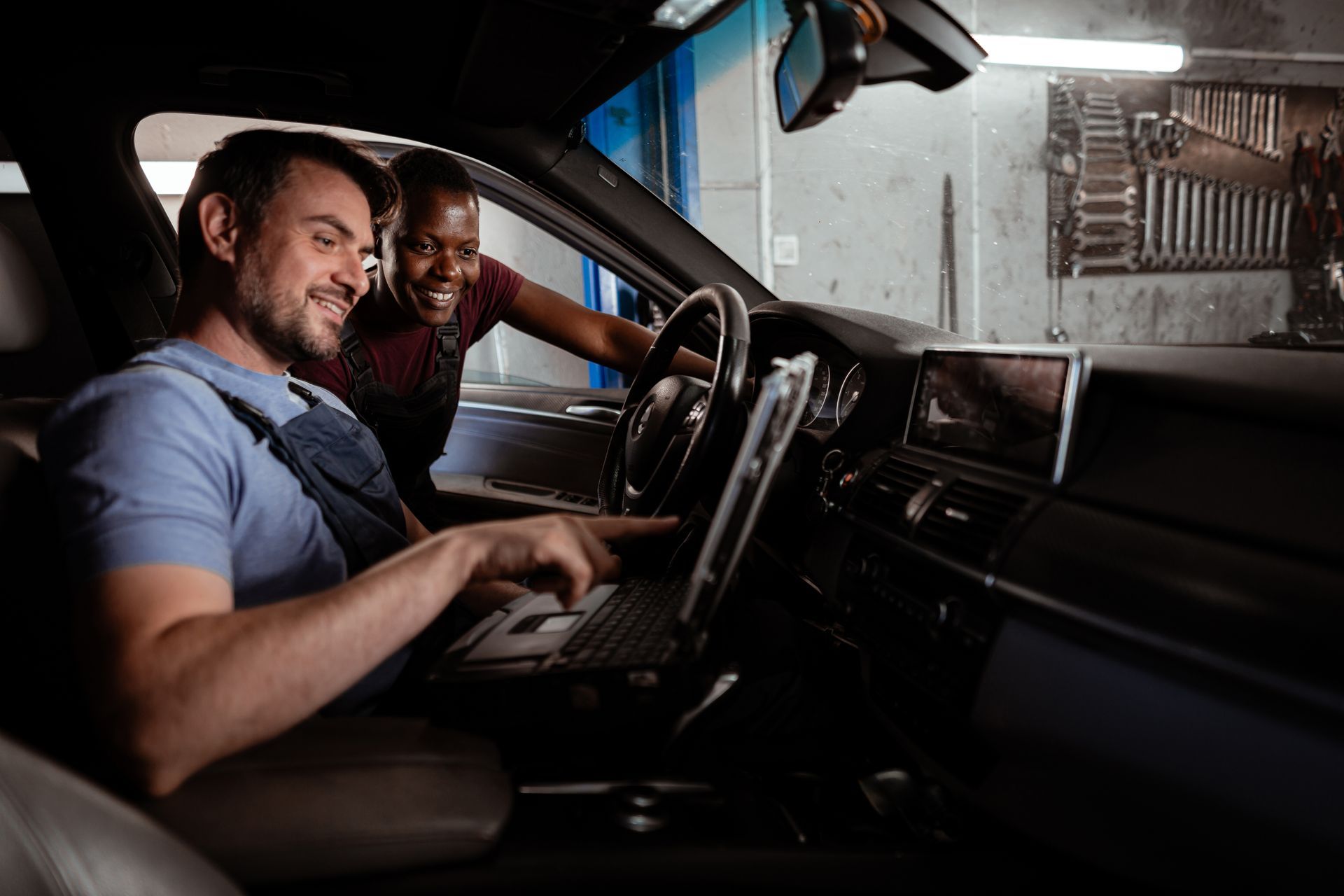 Two mechanics in a car; one points at a laptop on the dashboard, smiling; other mechanic smiles too. Garage setting.