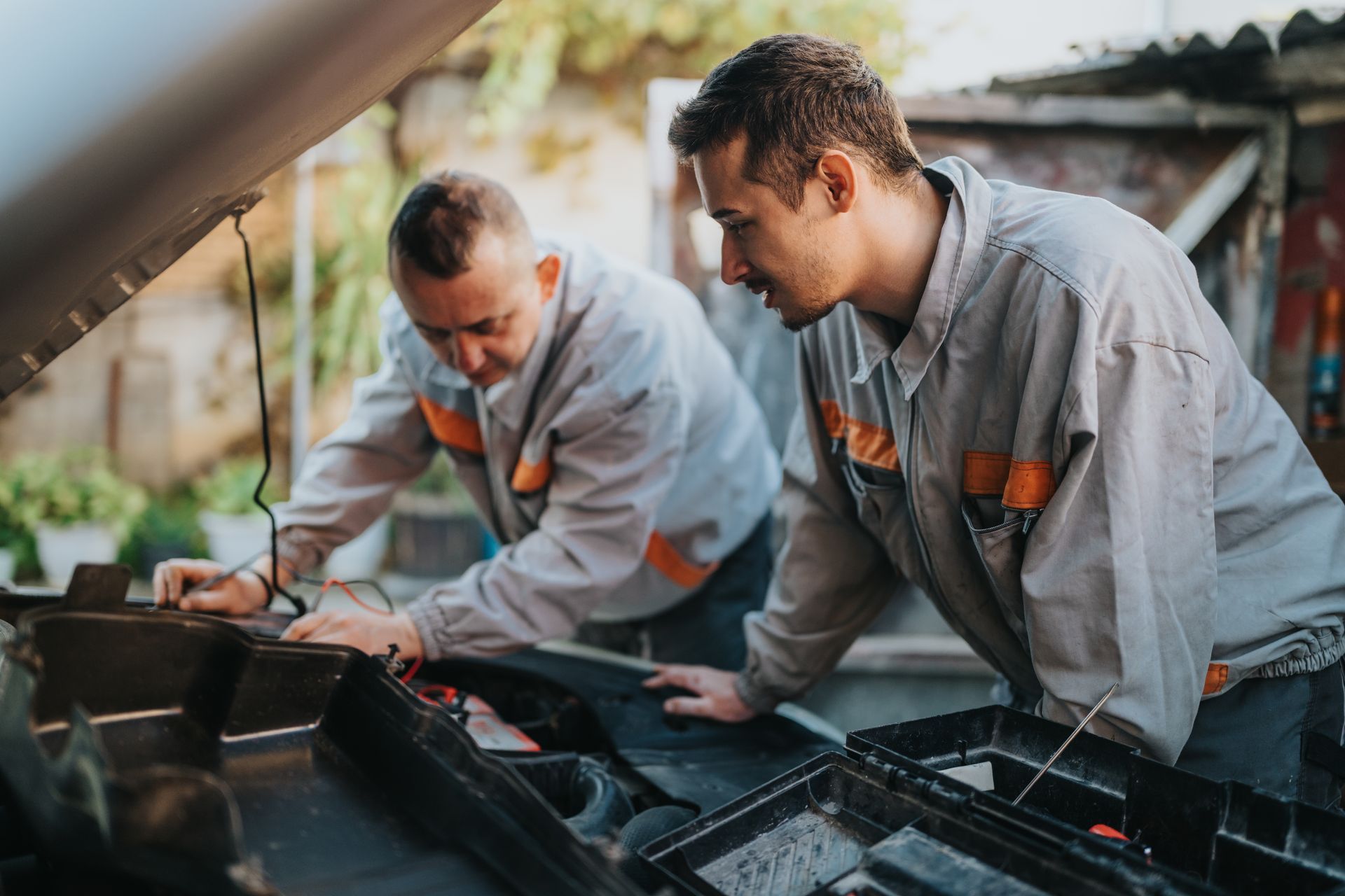 Two mechanics in gray uniforms inspect a car engine with the hood open, outdoors.