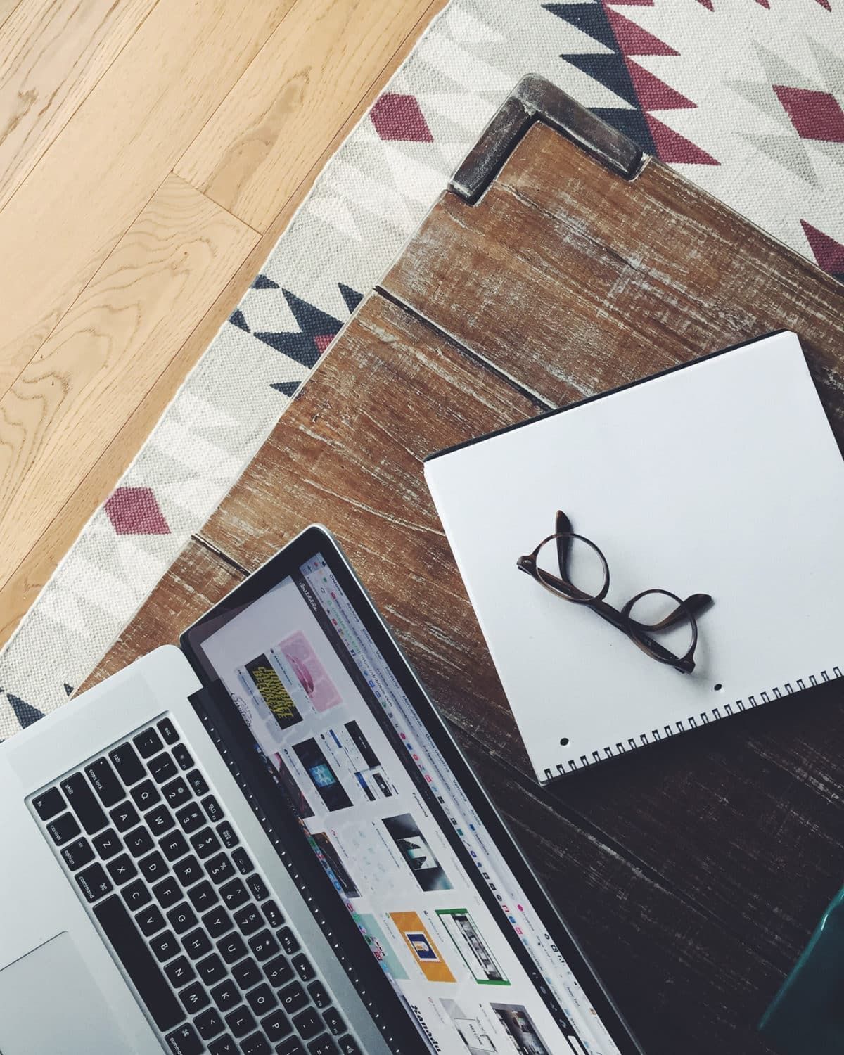 A laptop sits on a wooden table next to a notebook and a pair of glasses