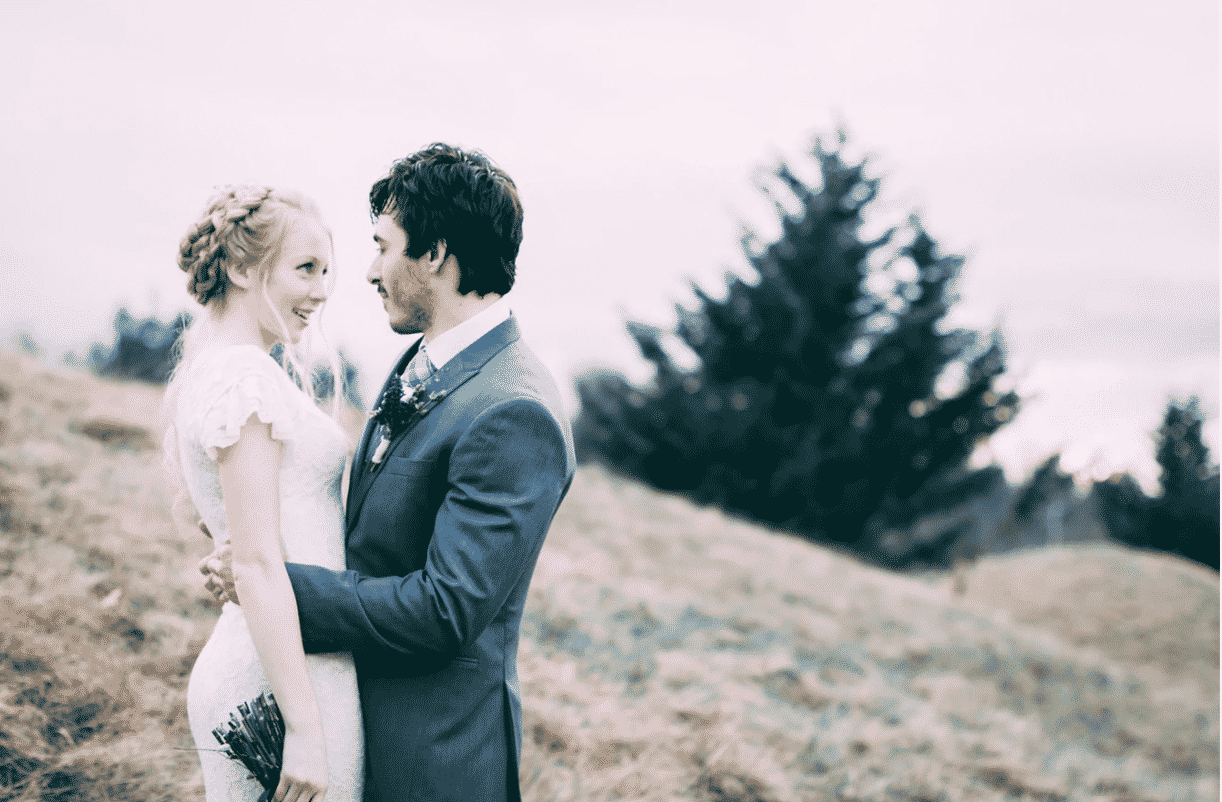 A bride and groom are standing on top of a hill looking at each other.