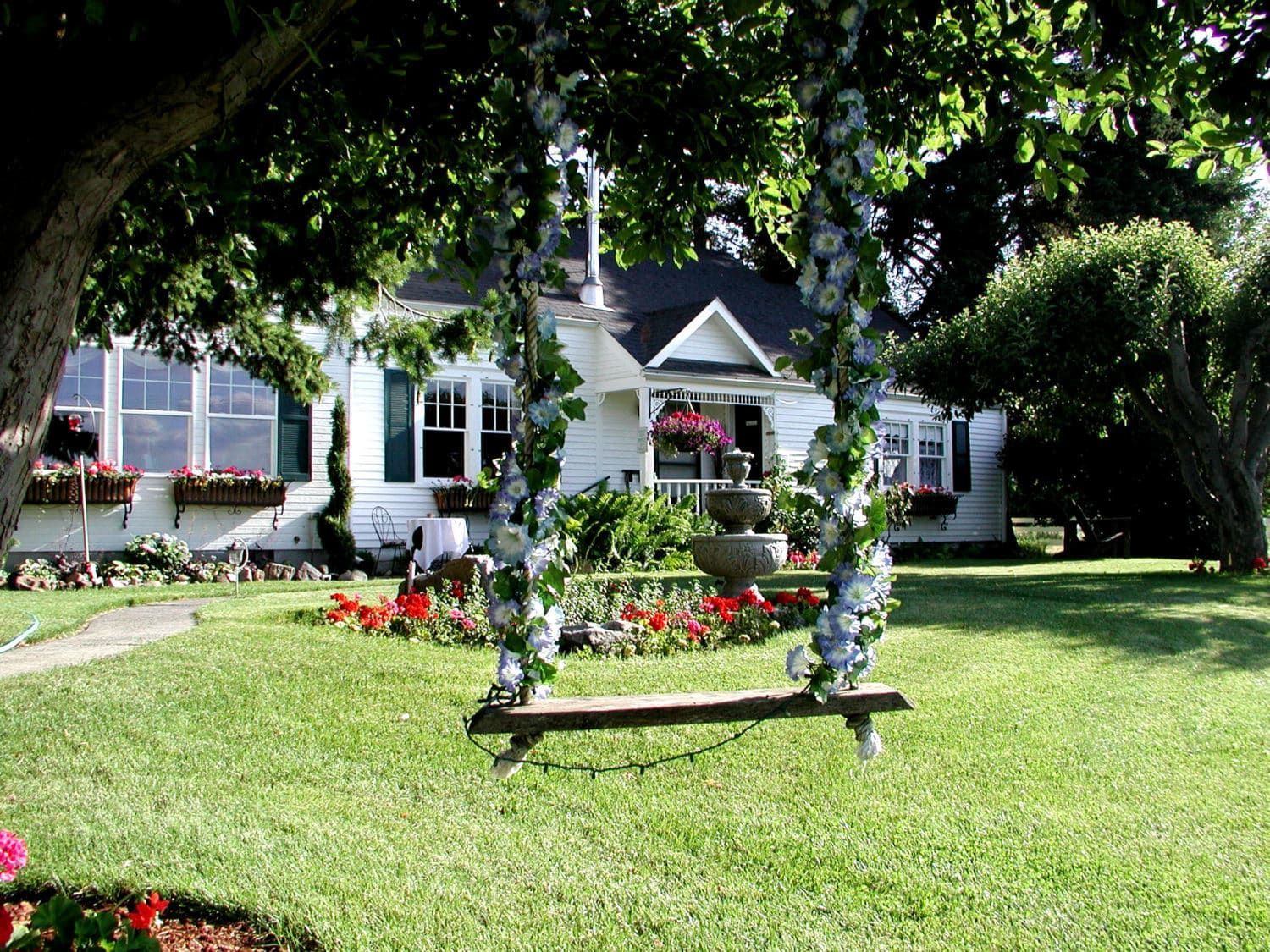 A wooden swing is hanging from a tree in front of a white house.