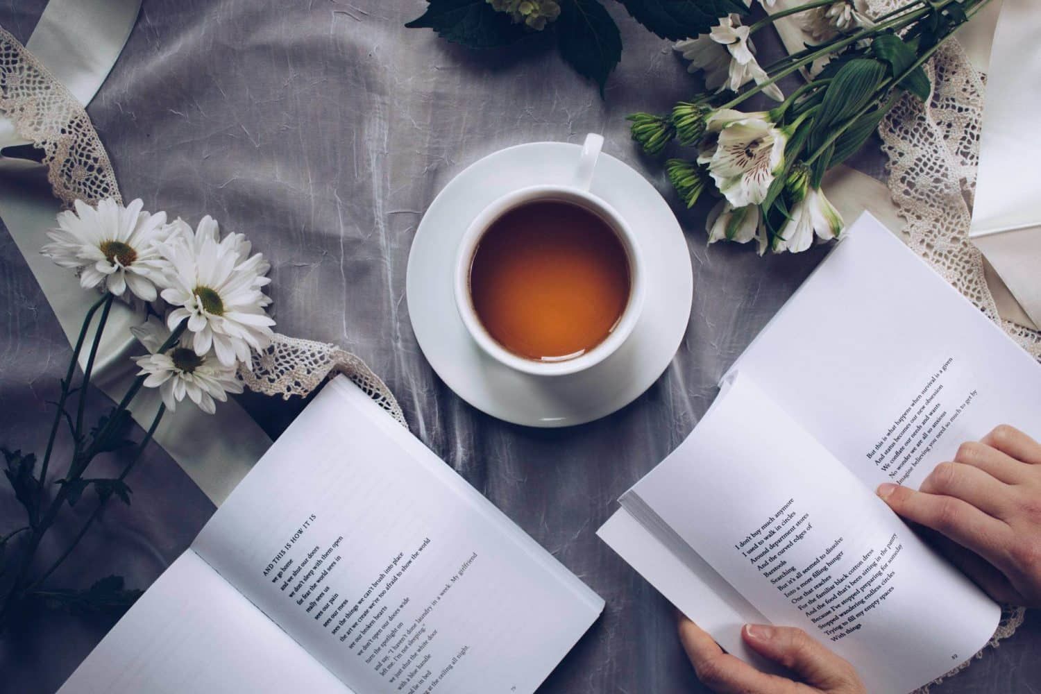 A person is reading a book next to a cup of tea and flowers.