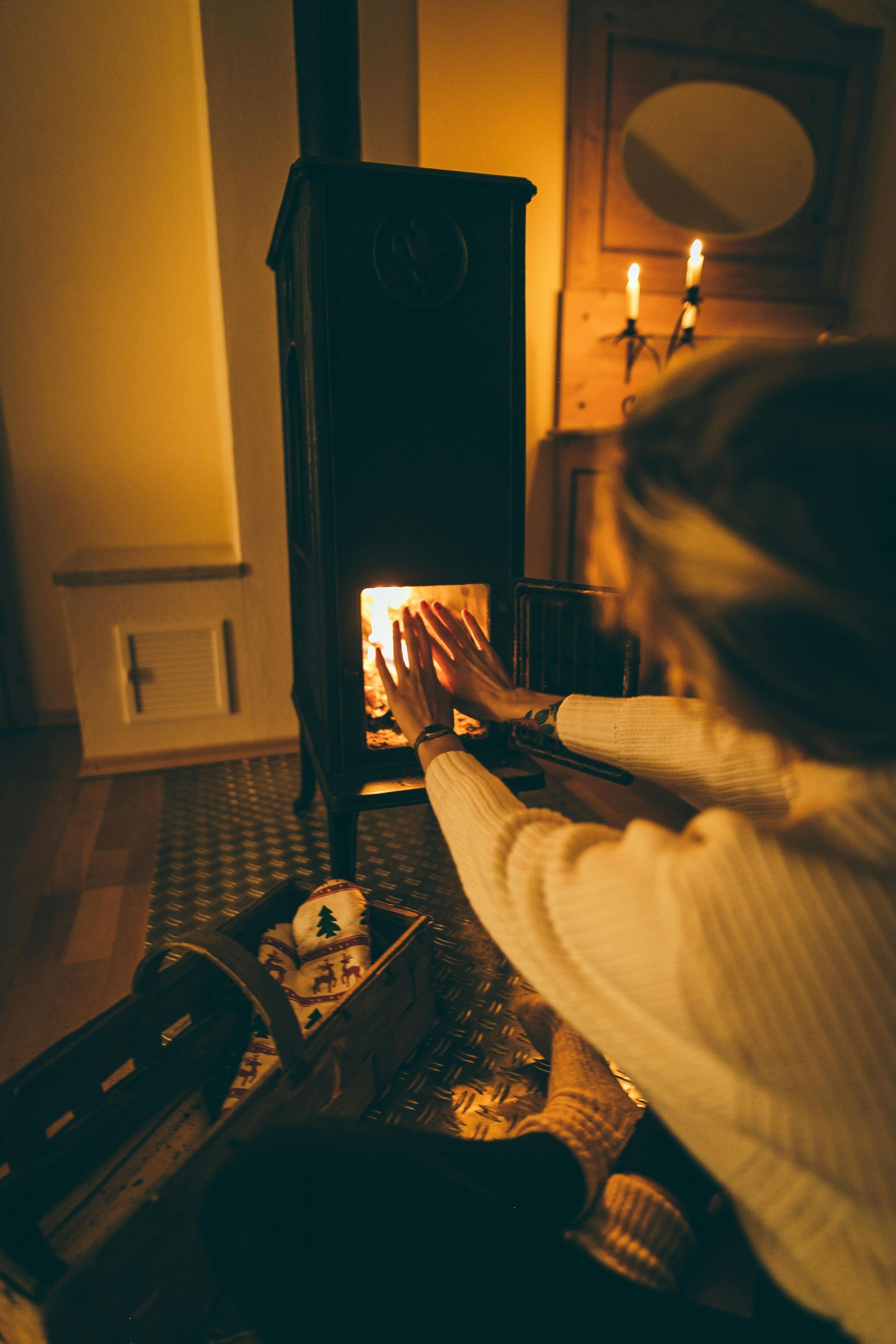A woman is sitting in front of a fireplace warming her hands.