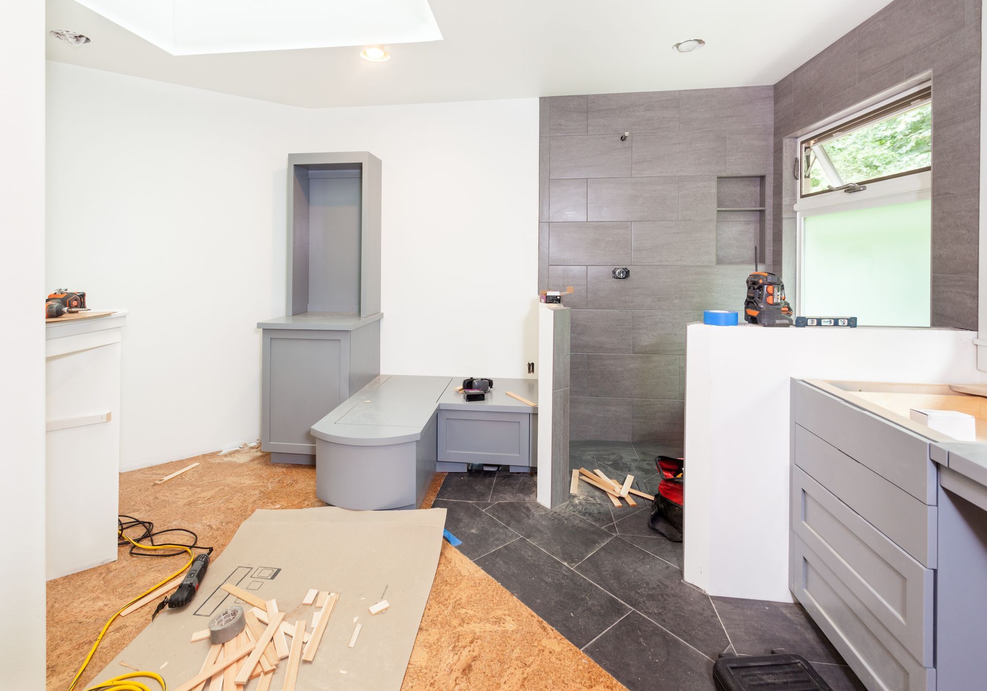 Bathroom under renovation; gray and white cabinets, gray tiled shower, exposed subfloor and construction debris.