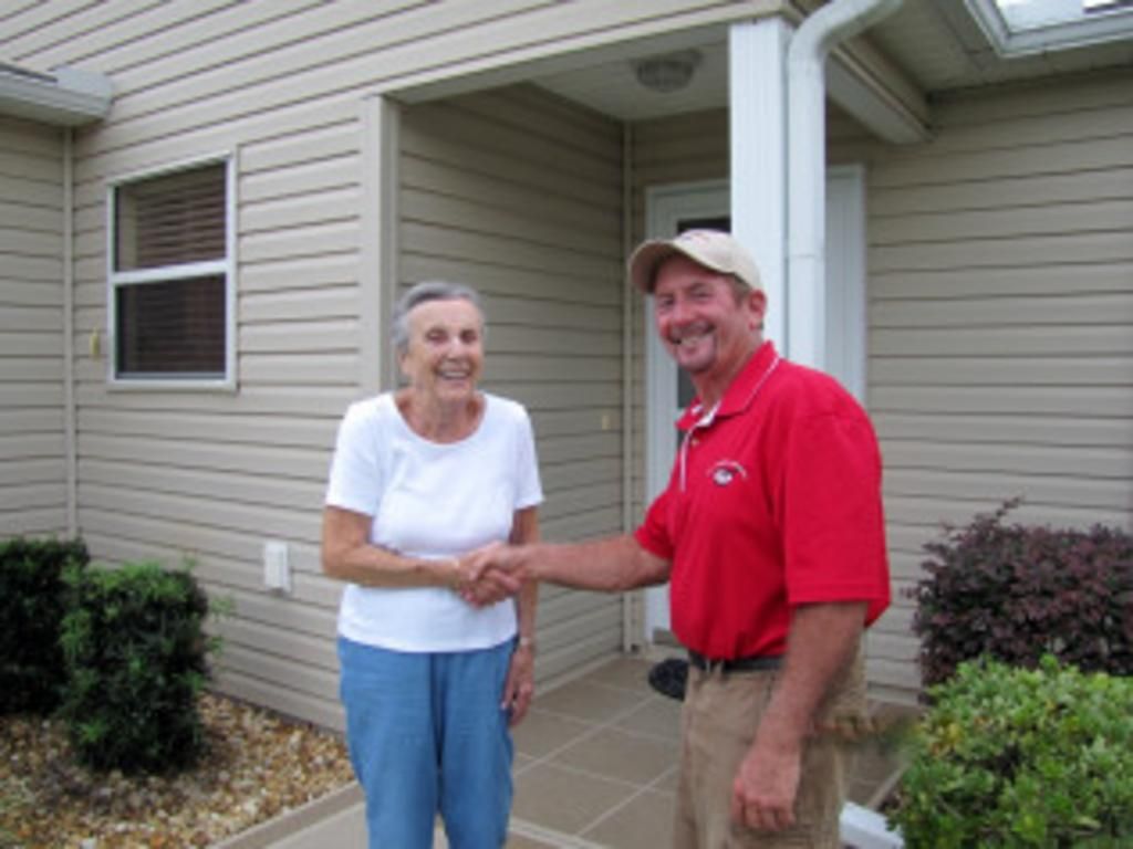 A man in a red shirt shakes hands with a woman in front of a house