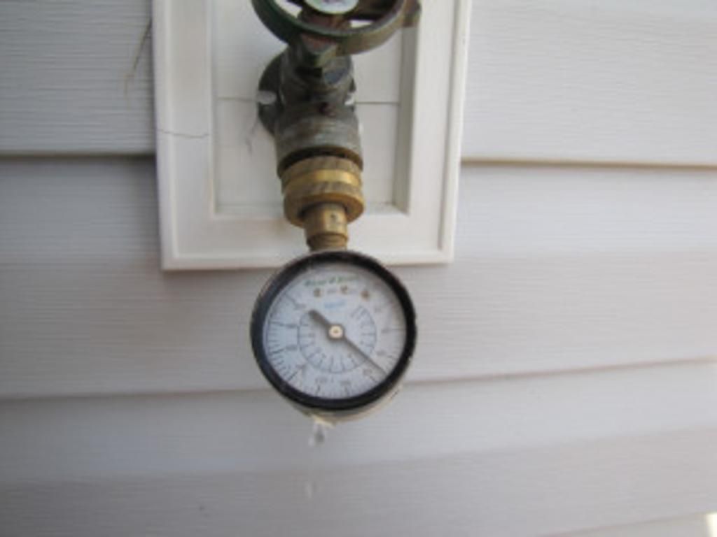 A pressure gauge is attached to a pipe on the side of a house.