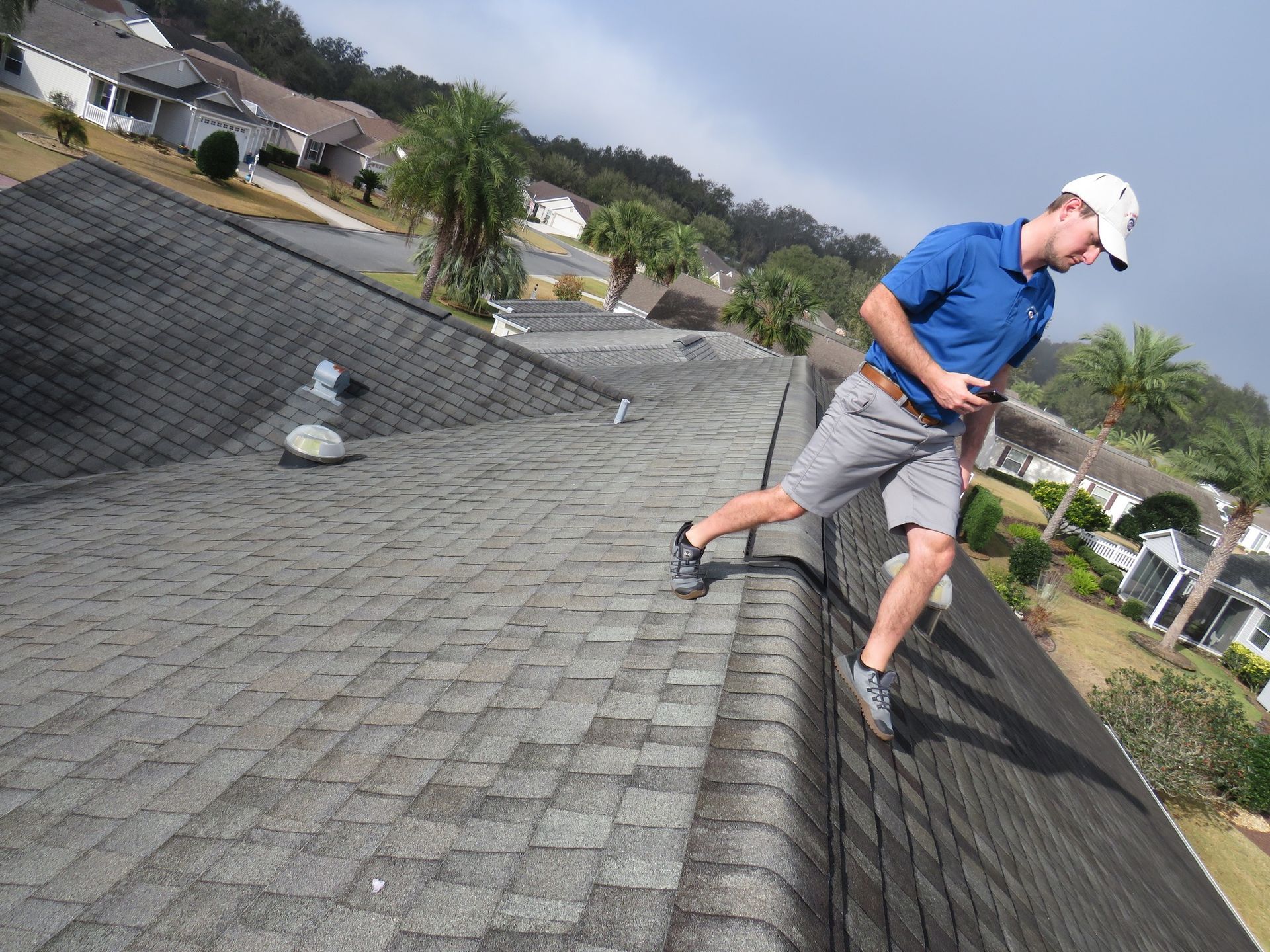 A person wearing a blue shirt and gray shorts walks along the ridge of a residential shingled roof on a sunny day.