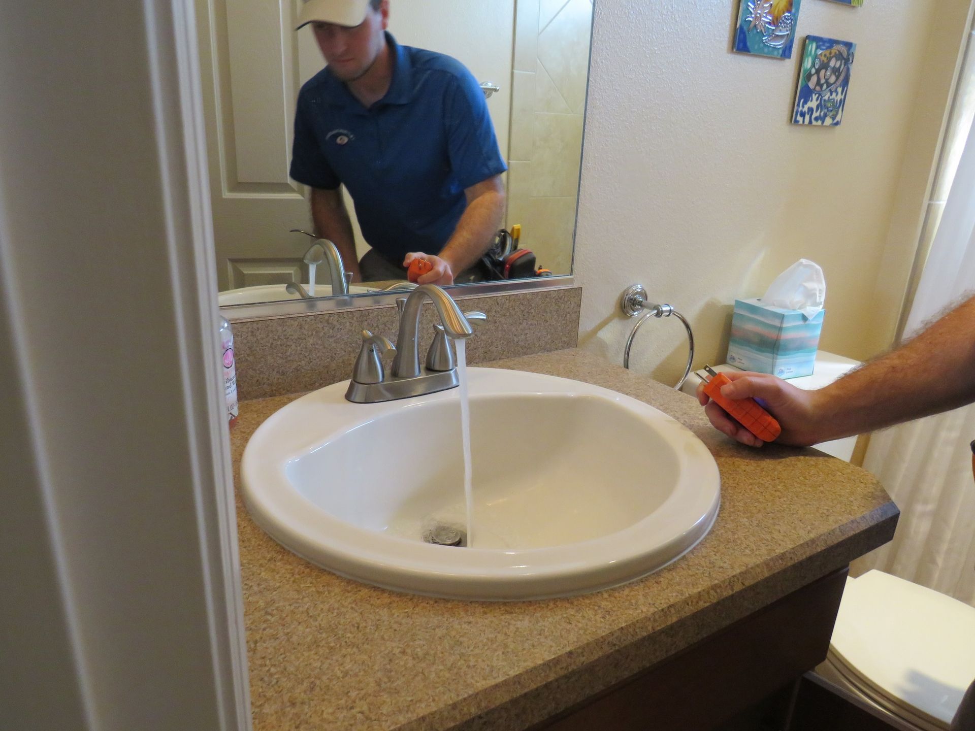 A person in a blue shirt holds a moisture meter near a bathroom sink with running water, checking for leaks.