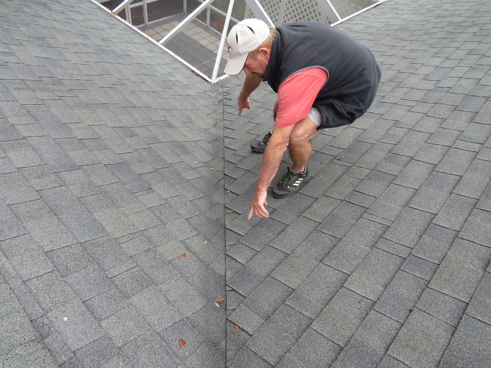 A person in a red shirt and dark vest crouches on a shingled roof, pointing at a seam where two roof sections meet.